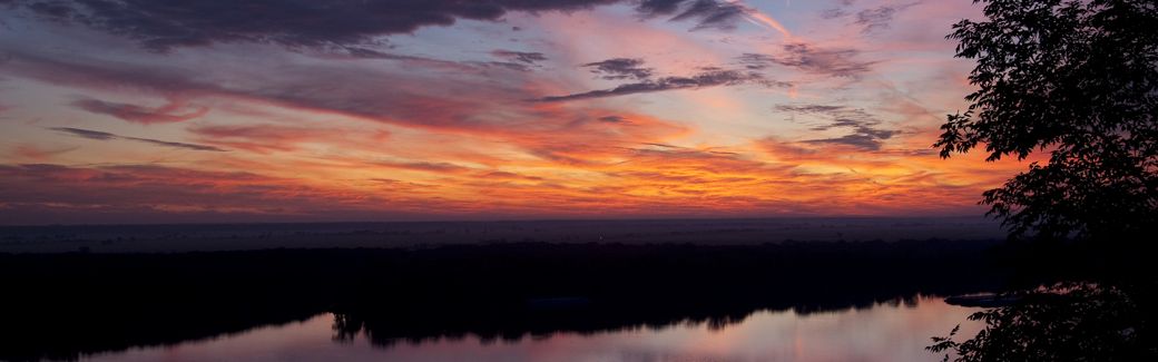 Sunrise illuminates wispy clouds with orange, pink, and purple hues over a still river in the foreground.