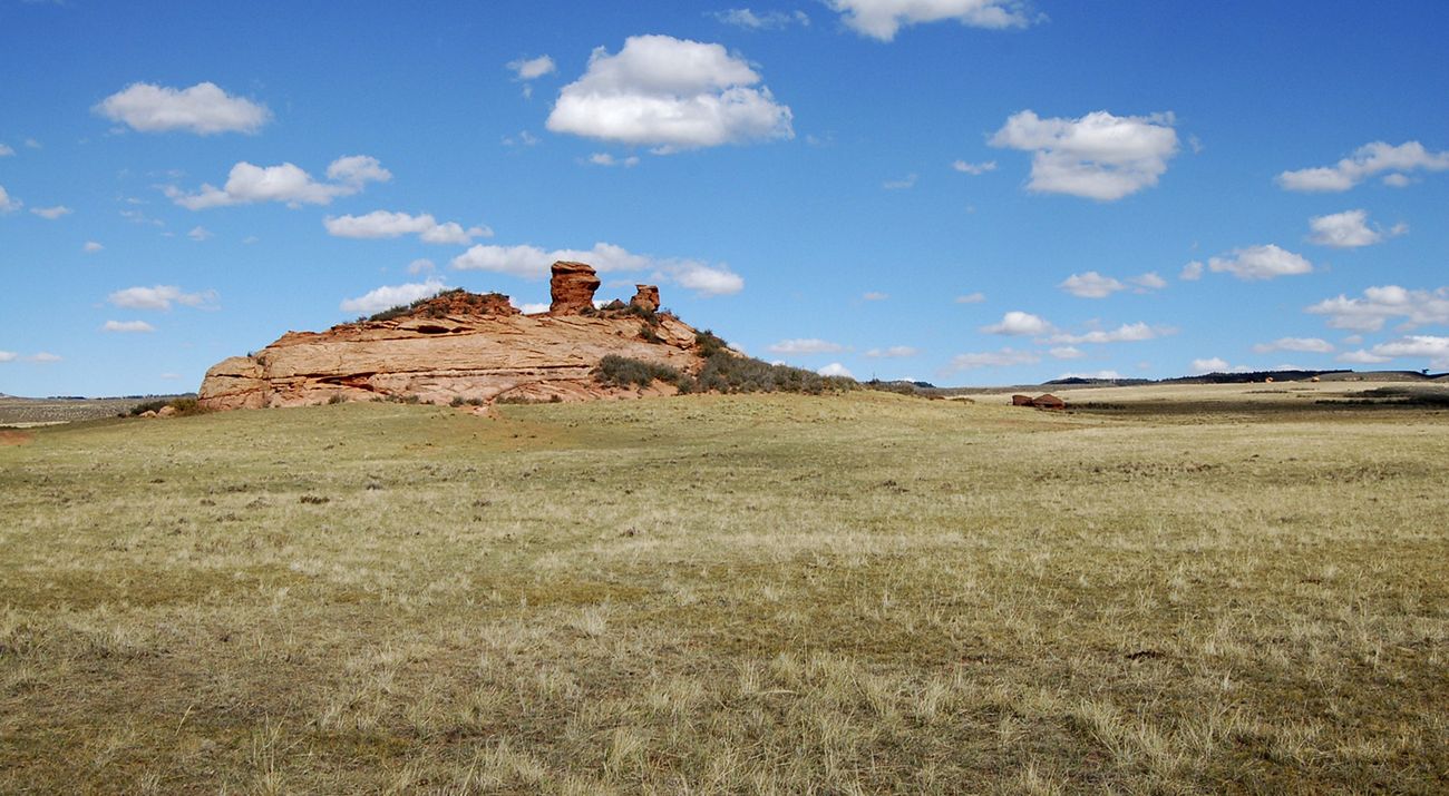 View across a dry grassland toward a red butte under a blue sky with patchy clouds.