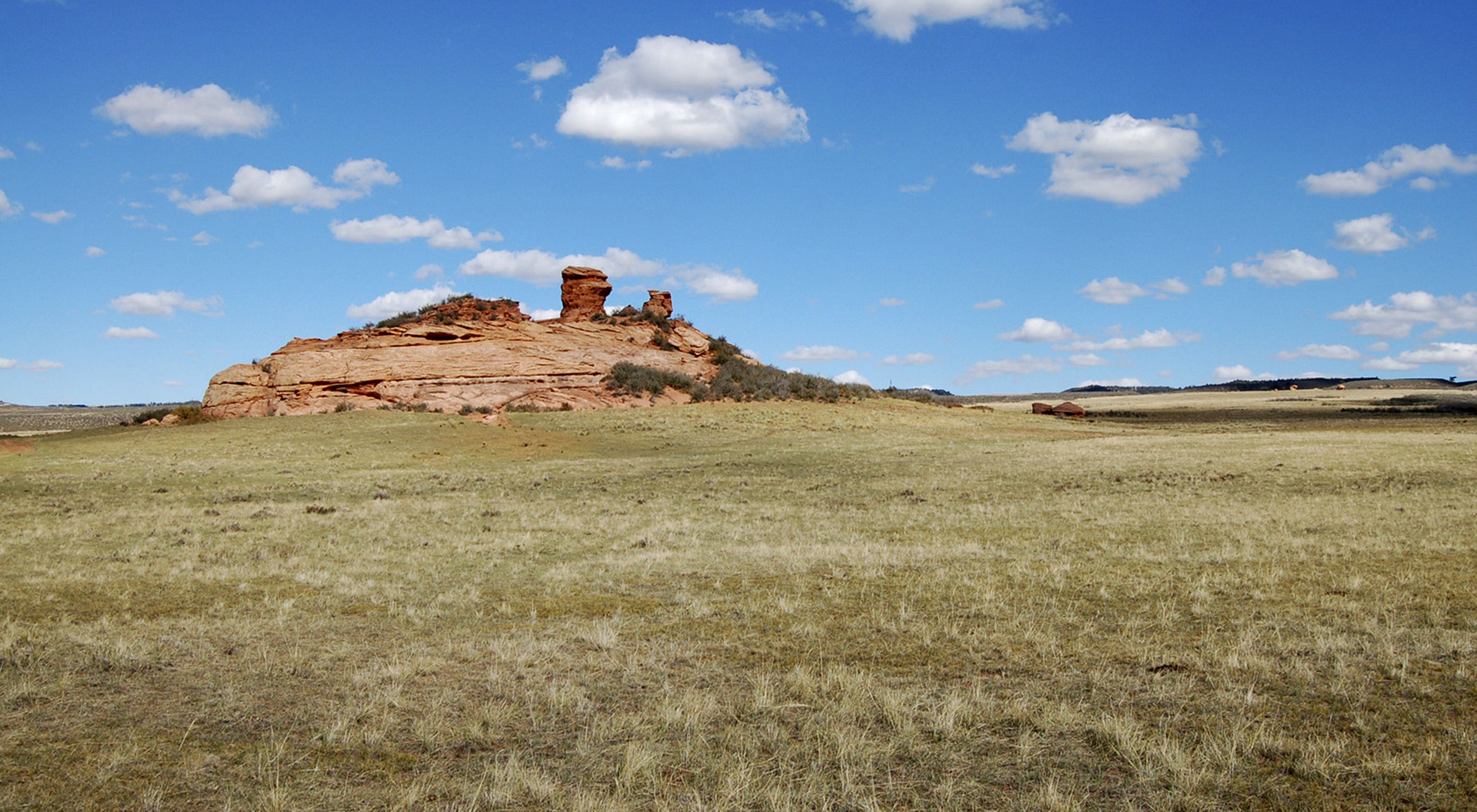View across a dry grassland toward a red butte under a blue sky with patchy clouds.