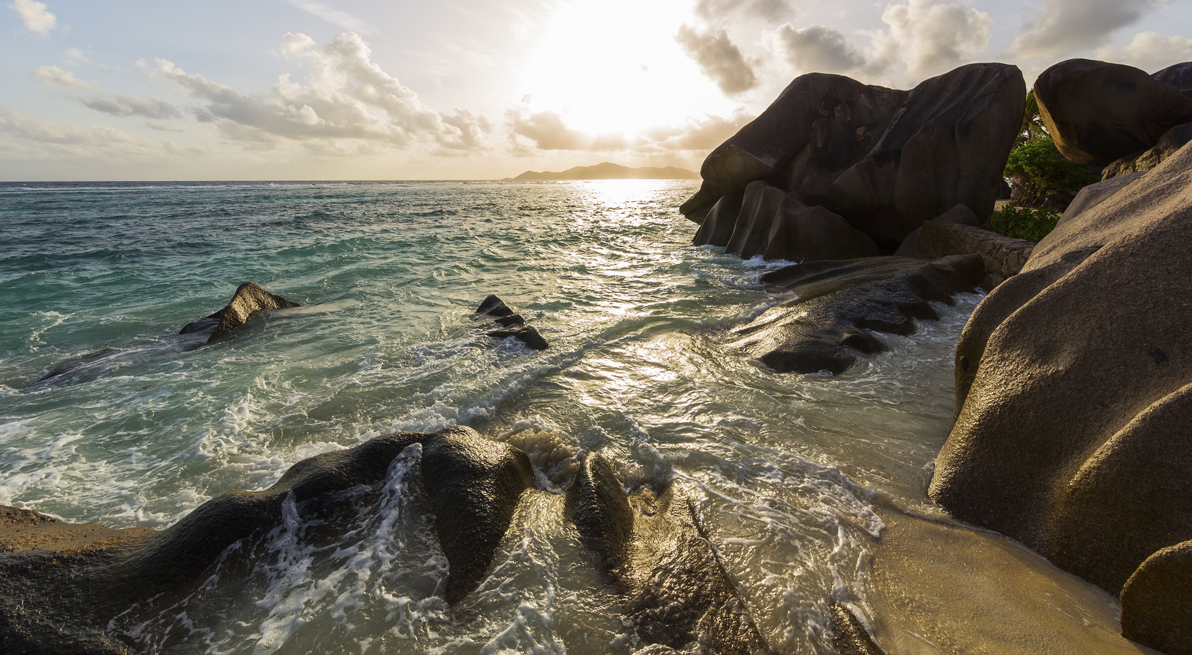 Granite boulders front turquoise ocean waves, under a blue, sunlit sky.