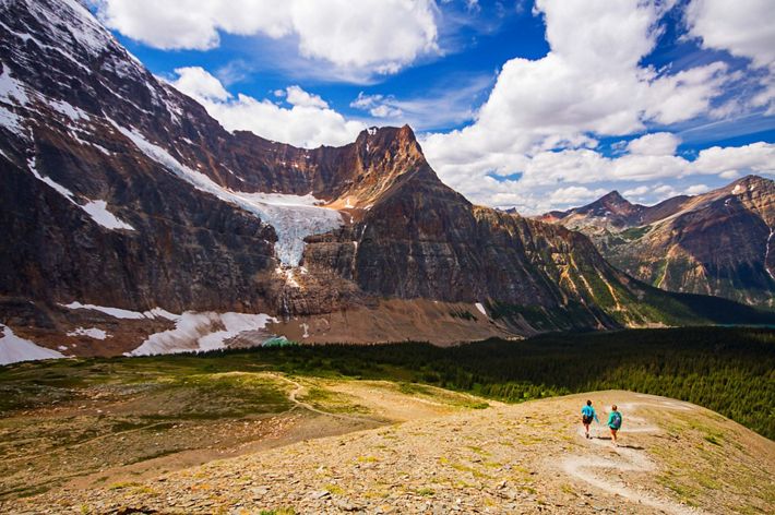 Two people walk on a trail in the direction of steep mountains with a forest at the base.