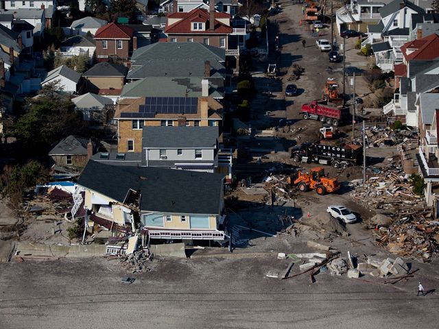 Aerial view of destruction caused by Hurricane Sandy. Rows of houses facing a beach show varying degrees of damage. In the foreground a yellow house is split in two.