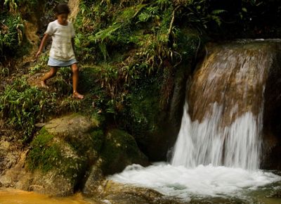 A young girl walks along the rocky edge next to a short waterfall.