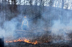 A women wearing yellow fire retardant gear monitors a small fire during a controlled burn. Smoke rises around her almost obscuring her from view.