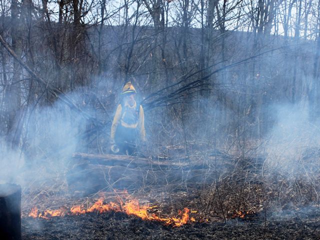 A women wearing yellow fire retardant gear monitors a small fire during a controlled burn. Smoke rises around her almost obscuring her from view.