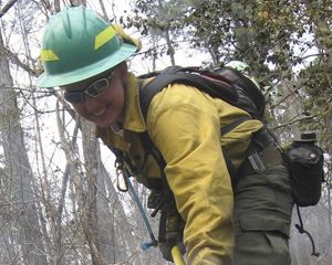 A woman in yellow fire gear smiles while she works outdoors.