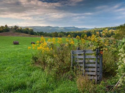 An open green pasture rolls into the distance before meeting a line of trees. A mountain range rises in the background topped by low hanging clouds. A small fenced enclosure is in the foreground.