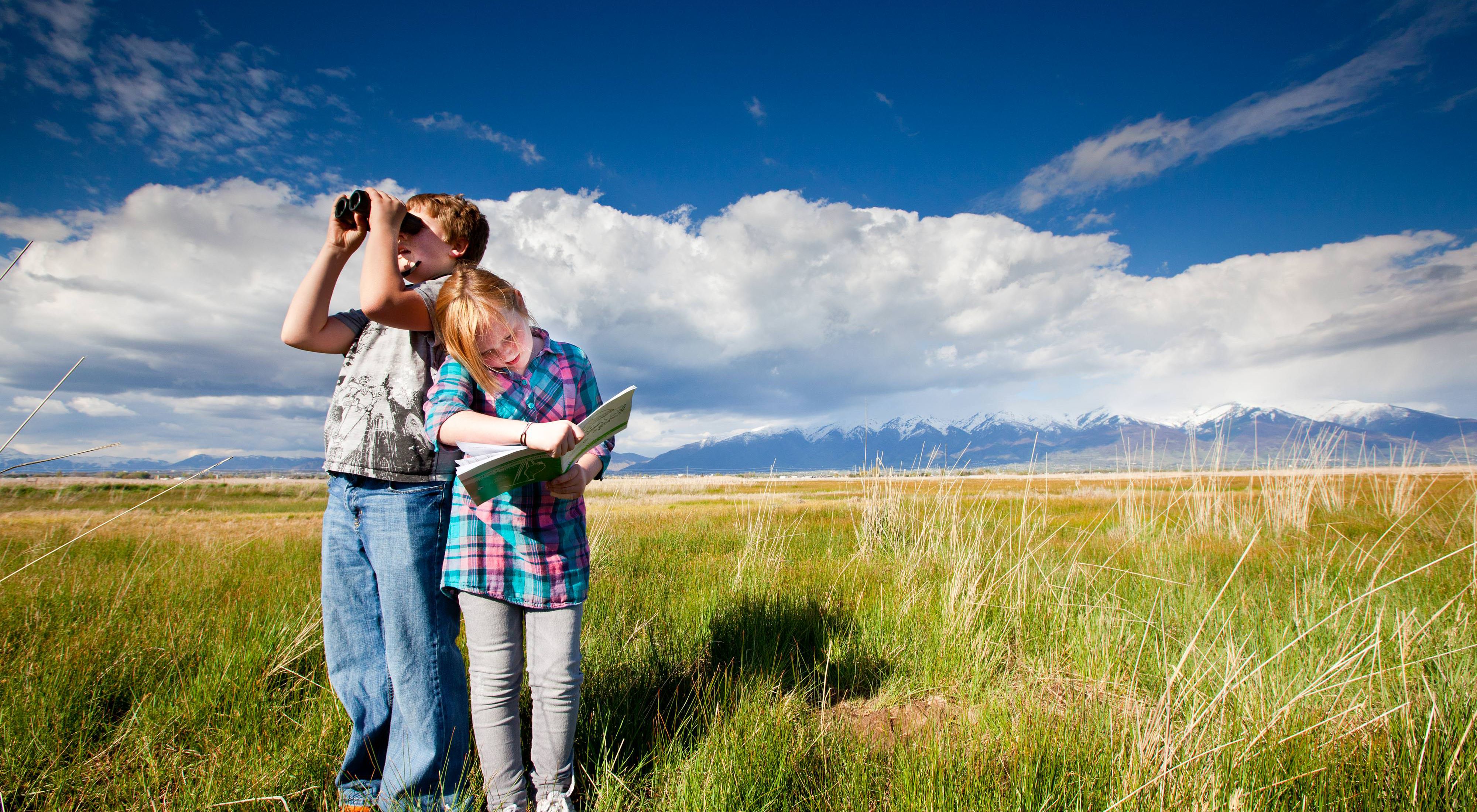 Two children stand in a grassy field with a snowy mountain range in the background. One child looks through binoculars and the other looks in a book.