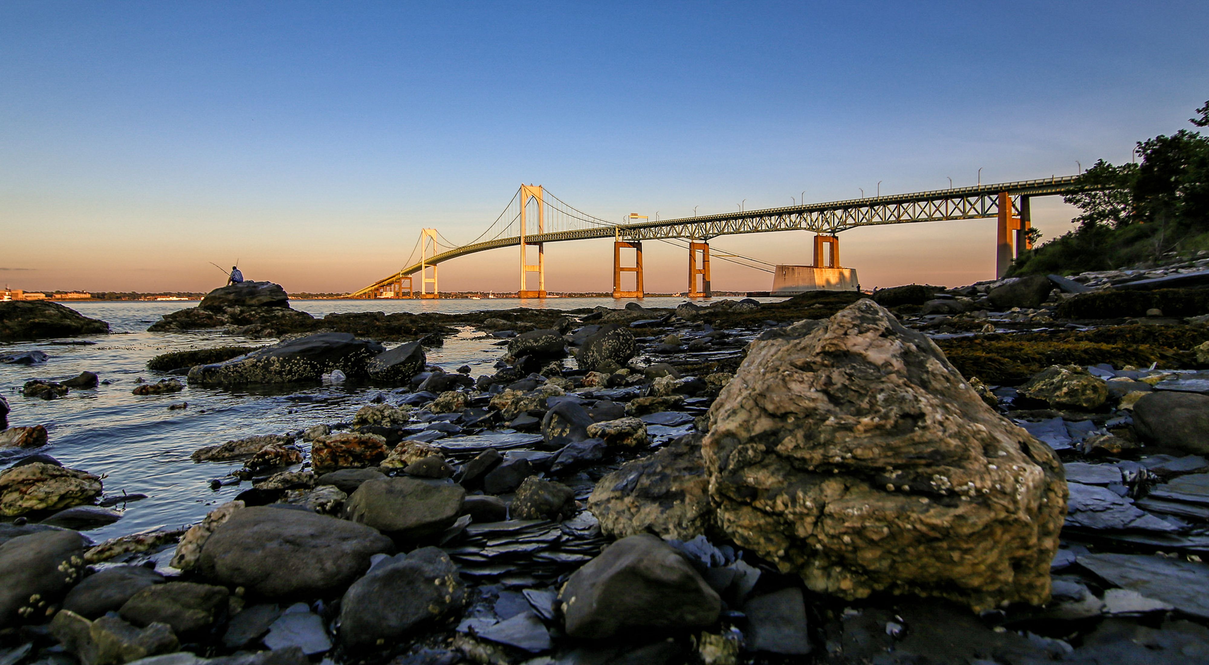 Rhode Island's Newport Bridge can be seen in the golden light of sunset beyond the rocky shoreline.