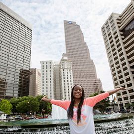 A young woman holds her arms out, with the Atlanta skyline behind her.