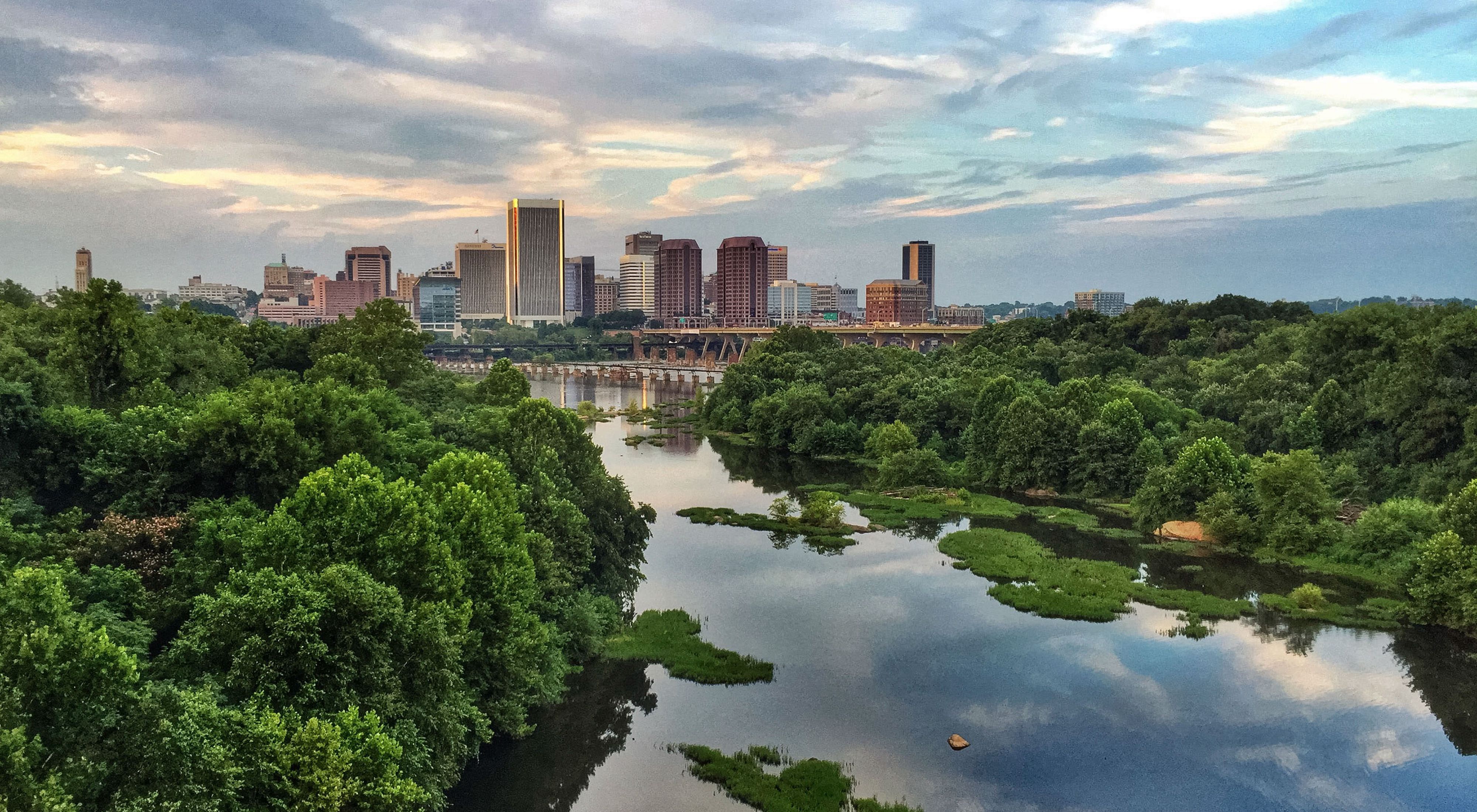 Aerial view of the James River looking downstream to downtown Richmond. A wide river flows between forested banks. An urban downtown of high-rise buildings is in the background.