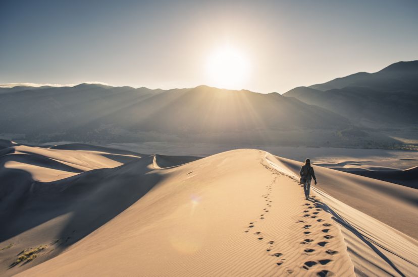 A lone figure walks along the crest of a tall sand dune, following a second set of footsteps. The sun hovers over the mountain range that lines the horizon.