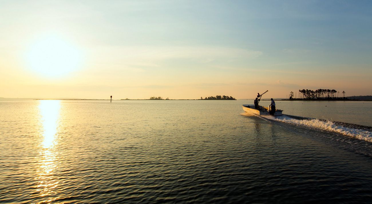 Two men in a flat bottomed jon boat motor into the open water of the Chesapeake Bay. The rising sun reflects off the calm water.