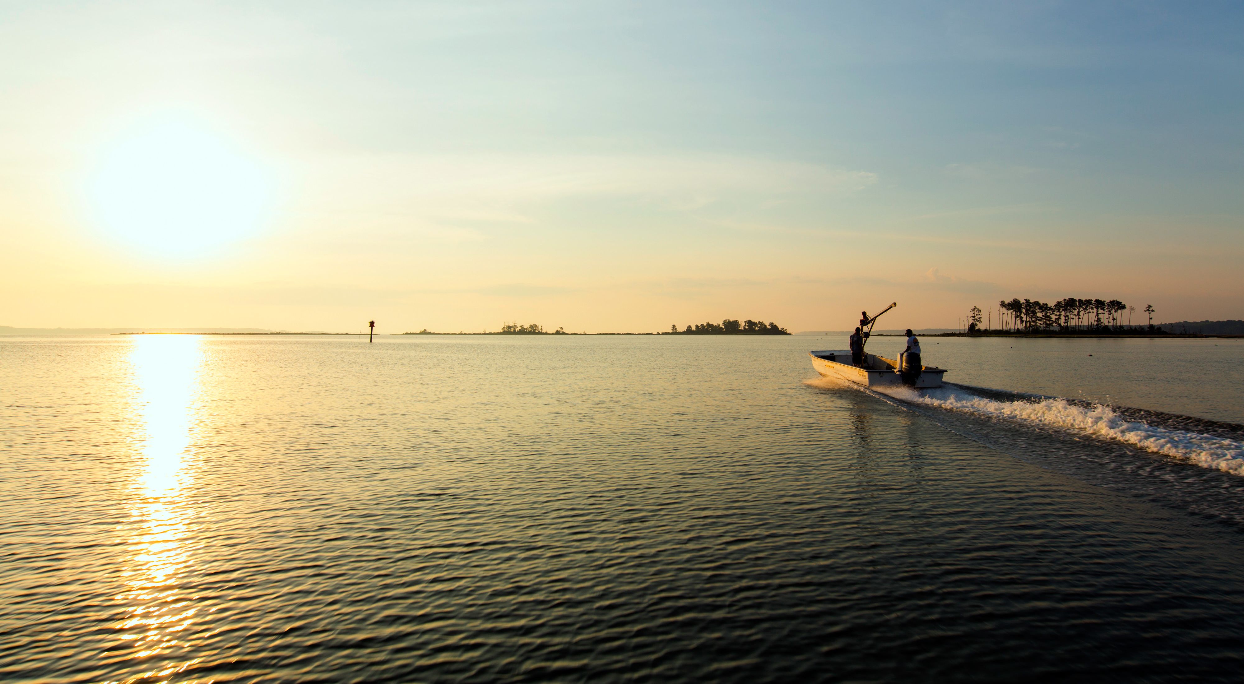 Two men in a flat bottomed jon boat motor into the open water of the Chesapeake Bay. The rising sun reflects off the calm water. 