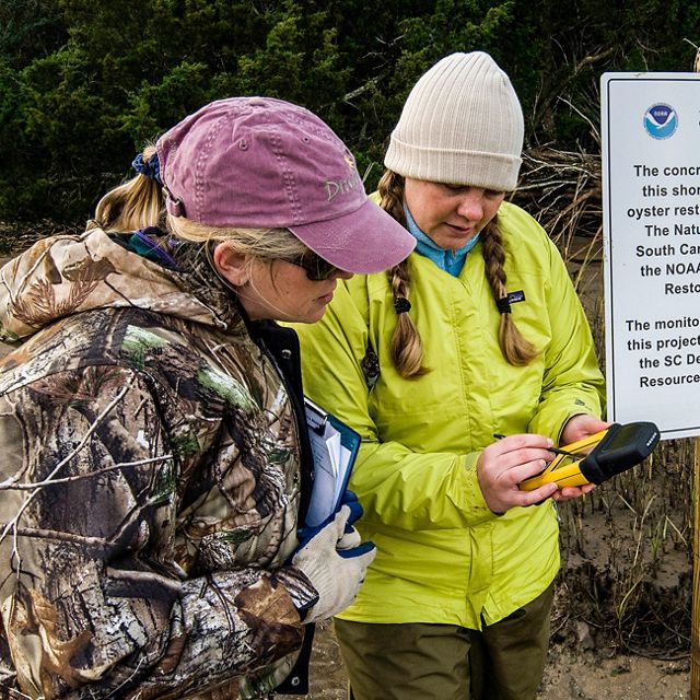 Two women look at a handheld GPS device while standing next to a TNC sign along a shoreline.
