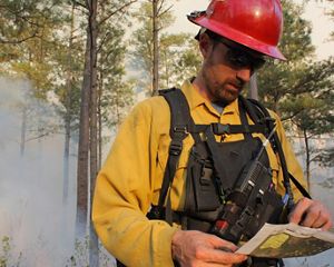 A man wearing yellow fire protective gear and a red helmet reviews a map during a controlled burn at Warm Springs Mountain. Thick smoke rises in the air behind him.