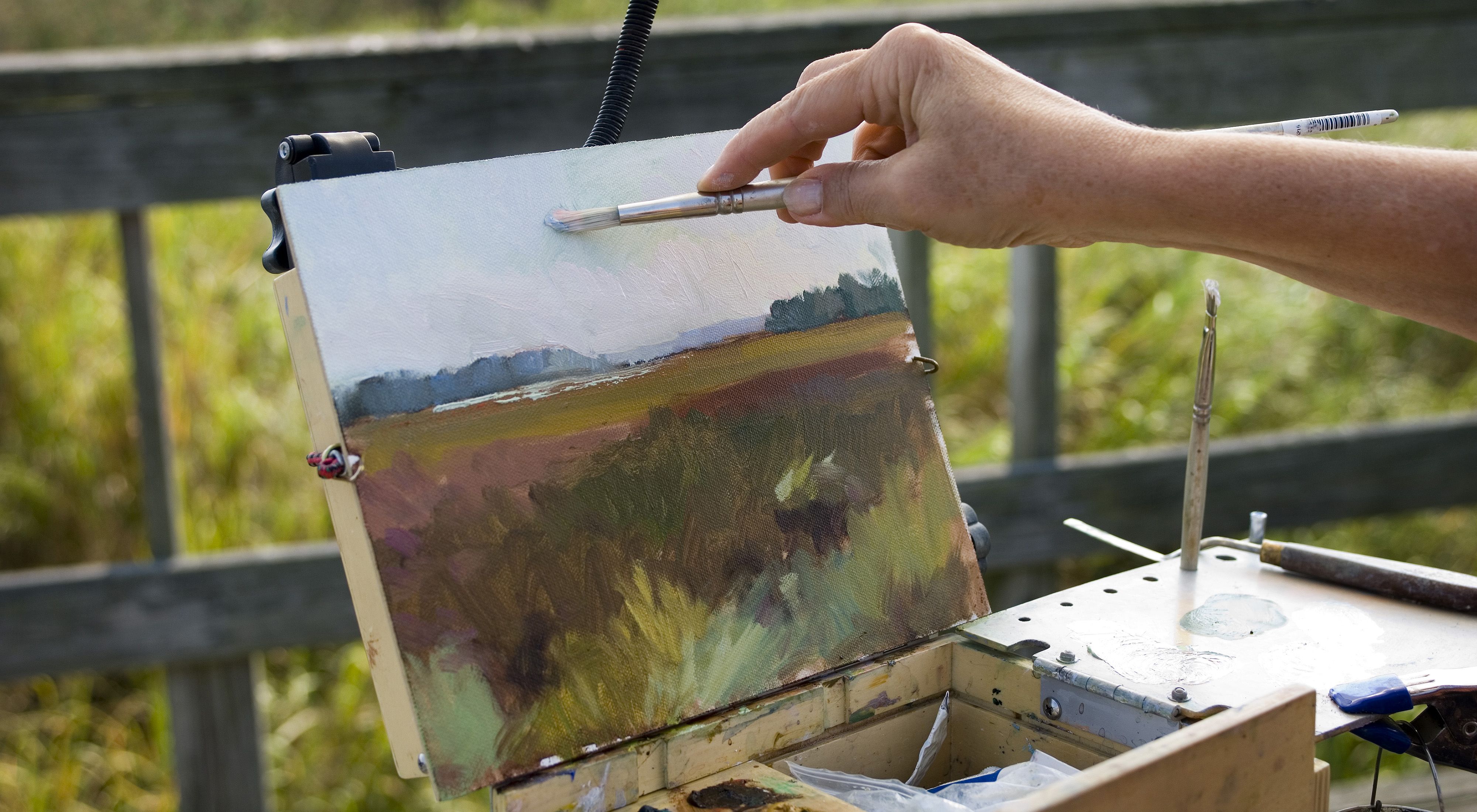 Close cropped view of a person holding a brush to a small canvas painting a wetland scene.