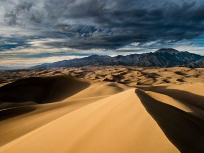 Sunlight and shadows create a wavelike appearance of a vast expanse of sand dunes with mountains in the distance.