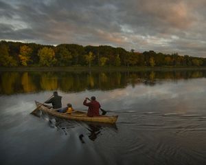 Anciano, adulto y niño reman una canoa en un río con arbustos y el cielo nublado en el fondo.