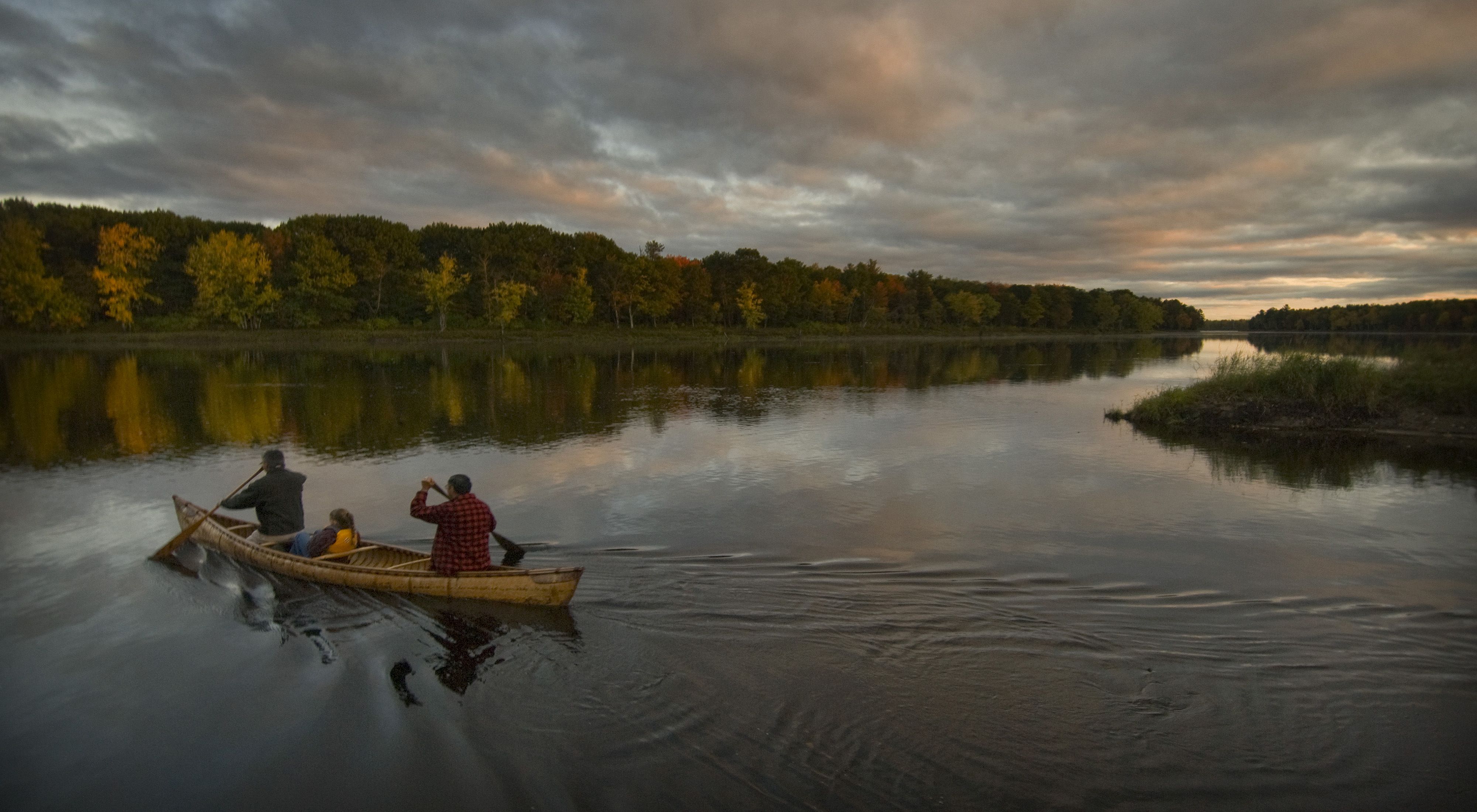 A family paddles across the Penobscot River in a birch canoe at sunset.