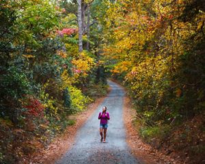 A smiling woman walks along a gravel trail surrounded by fall trees.