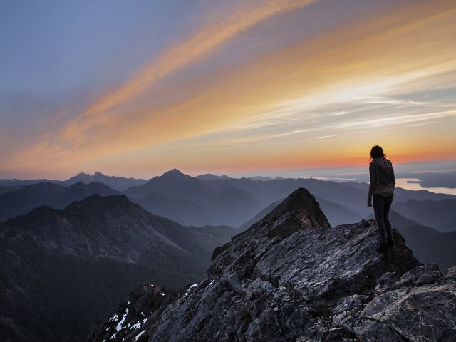 A hiker stands on a rocky outcrop looking out over mist shrouded peaks that stretch to the horizon.