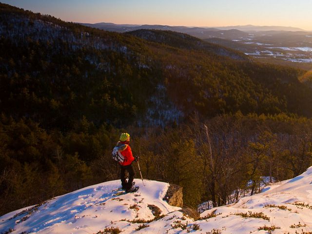 A person stands on a snow dusted overlook, looking out at the shadowy valley below.