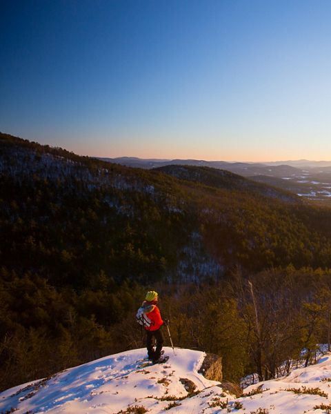 A snowshoer stands at the top of a snowy mountain overlook.