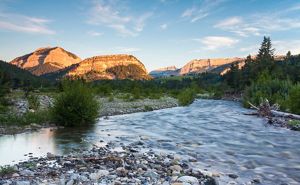 A clear mountain stream flows through a rocky riverbed bordered by evergreen trees, with sunlit cliffs and rugged peaks rising in the background under a blue sky.