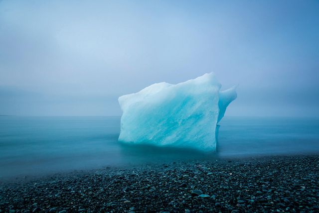 A portion of an glacier sits at the shoreline of a black sand beach under an overcast sky.