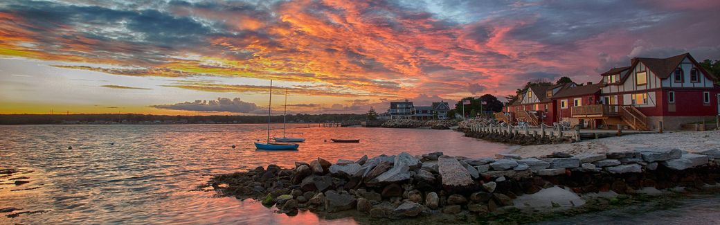 Sunset illuminates clouds with spectacular hues of orange, pink, and purple, with a tranquil waterfront and village in the foreground.