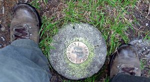 Looking down between two feet in brown boots at a stone border pin with a bronze marker that says United States on one side and Canada on the other.