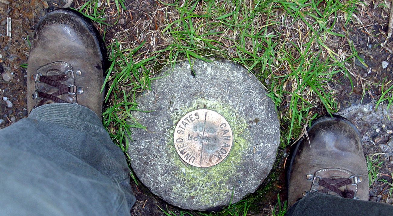 Looking down between two feet in brown boots at a stone border pin with a bronze marker that says United States on one side and Canada on the other.