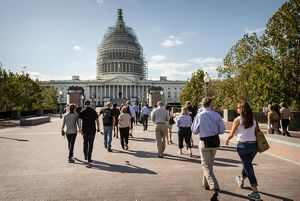 A large group of a dozen people walk alone the brick paved plaza towards the US Capitol building in Washington, DC. The building's dome is shrouded in scaffolding.