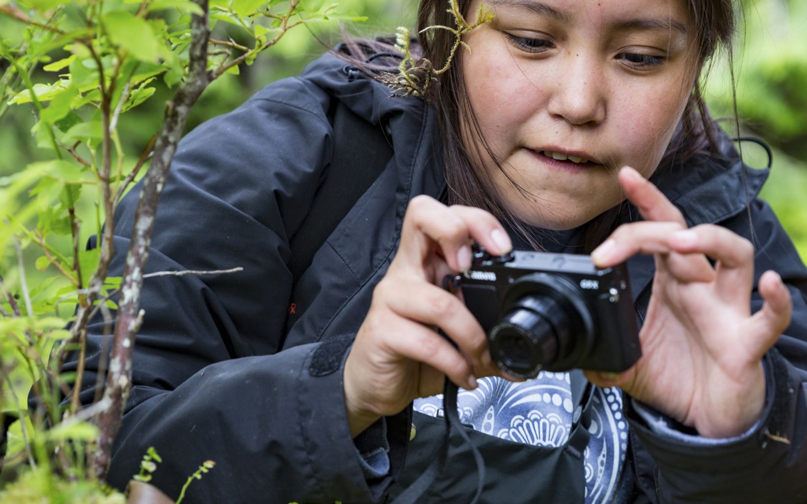 a girl with black hair points a small black camera at a leaf to take a picture
