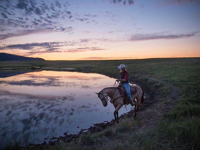 A woman on horseback stands next to a body of water in a field at sunset.