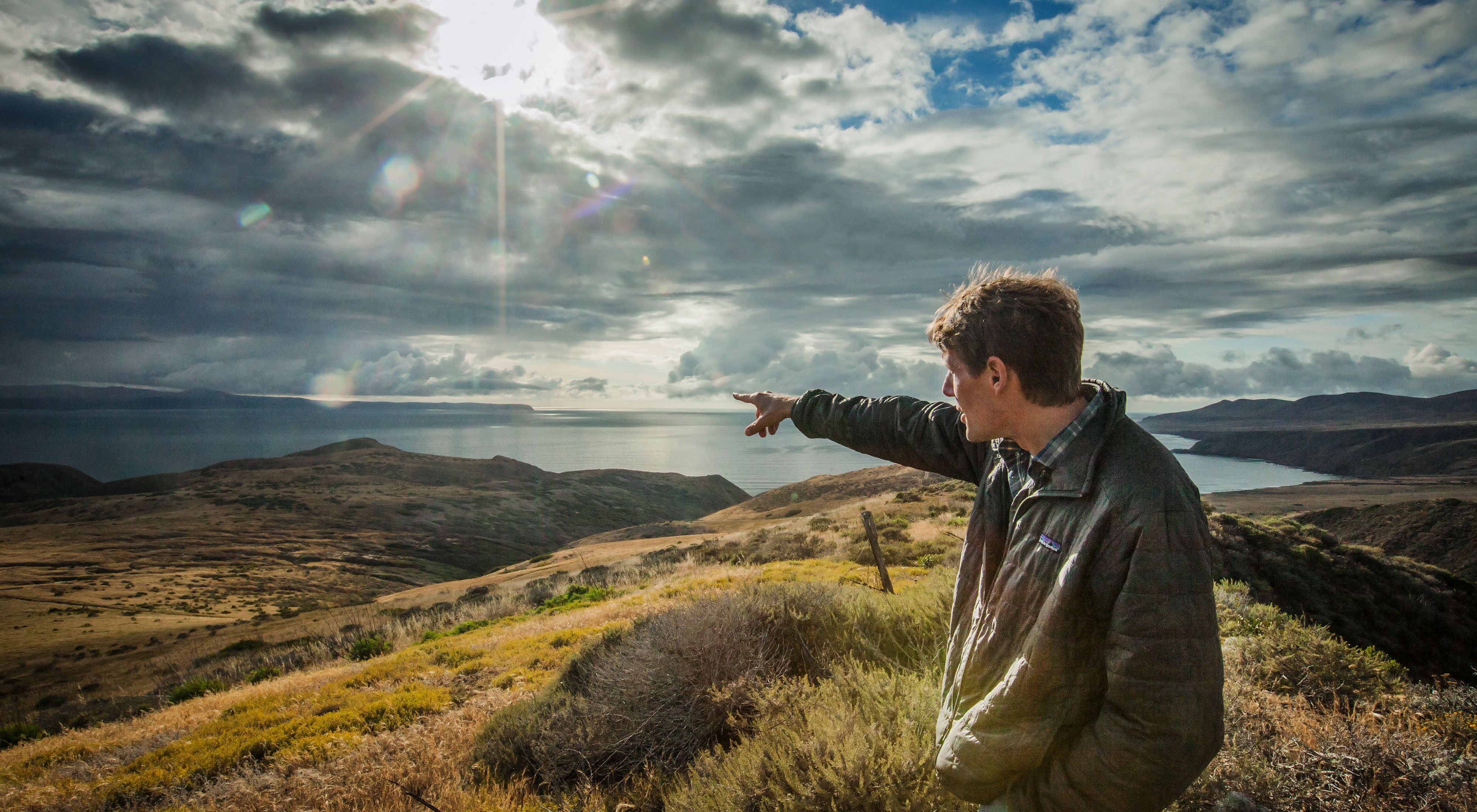 A man stands on a bluff at sunset and points out towards the ocean over grassy hills.