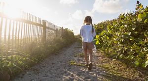 A child walks along a trail next to a sand dune and coastal foliage.