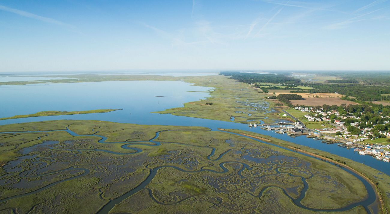 Aerial view of Watchapreague, VA. Meandering channels through coastal marsh, leading to a wider body of water, and a small coastal town with piers and buildings.