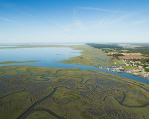 Aerial view of Watchapreague, VA. Meandering channels through coastal marsh, leading to a wider body of water, and a small coastal town with piers and buildings.