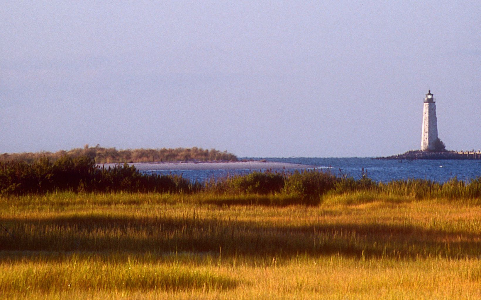 The New Point Comfort lighthouse rises from a spit of land jutting into the Chesapeake Bay. An open area of short, yellow winter grass is in the foreground.