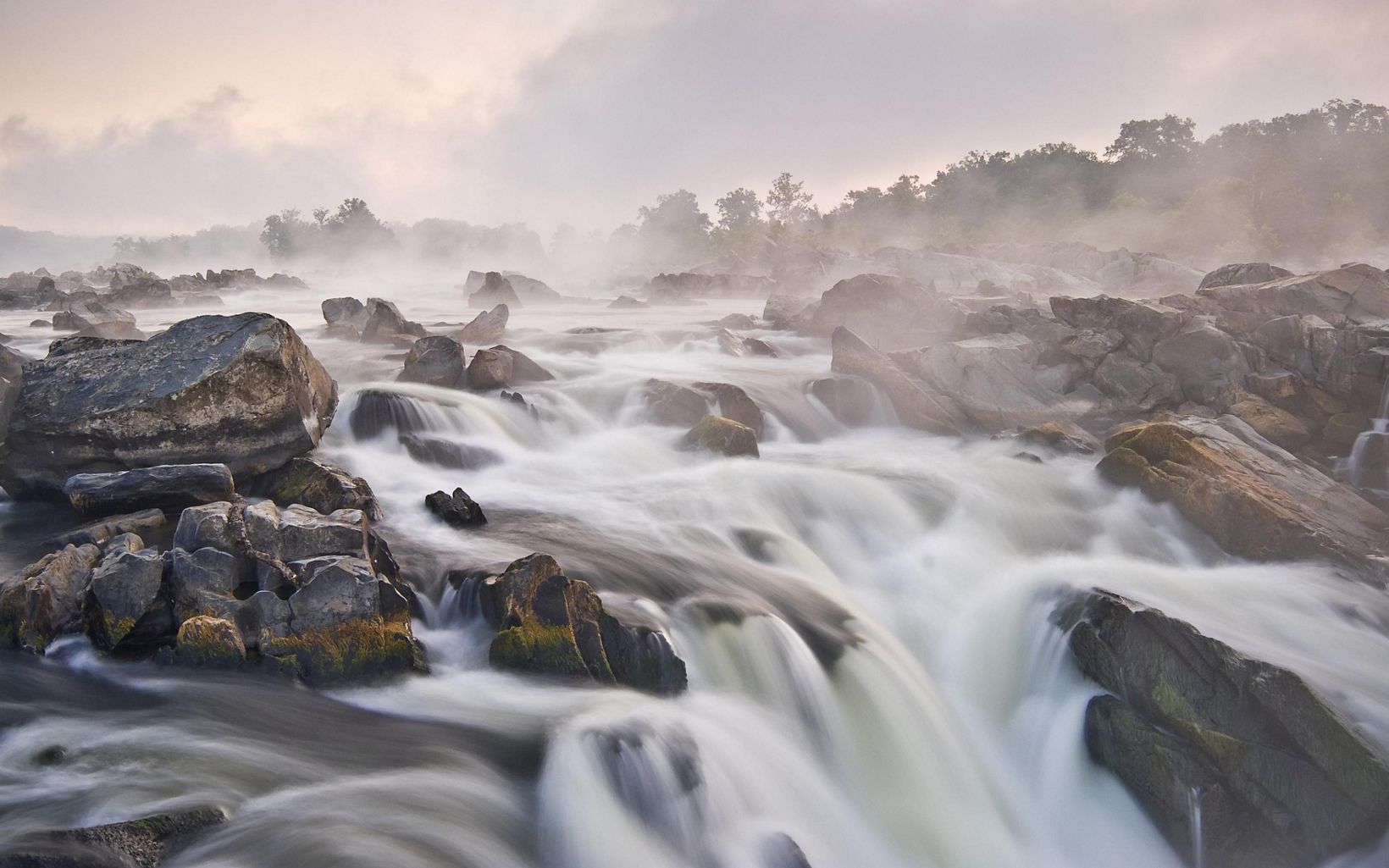 White water rushes over large rocks to create a series of short falls on the Potomac River. Fog rises from the water in the early morning obscuring the tree lined banks.
