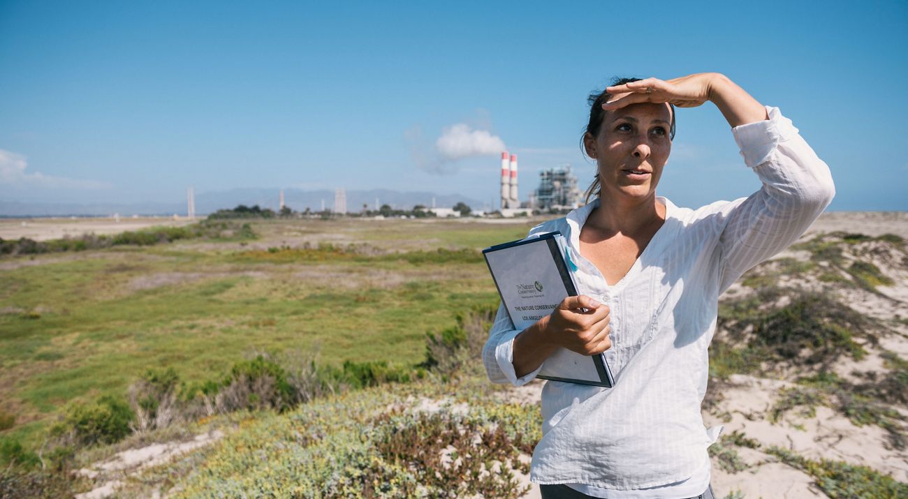 A woman holding a binder stands in a field and looks out in the distance while using her hand to shield her face from the sun.