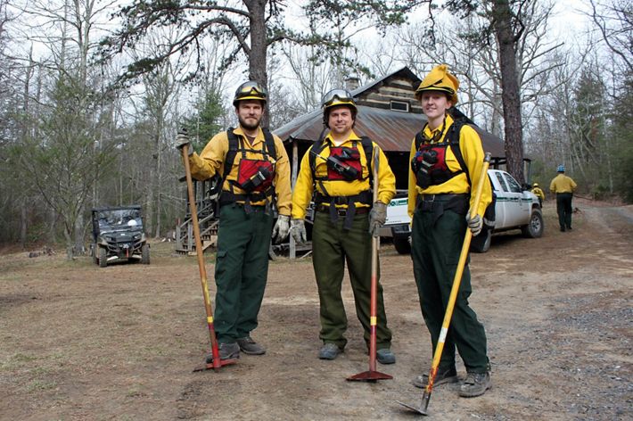 Three people pose together in front of a rustic cabin. They are wearing yellow fire retardant gear and holding long handled hoes. A white pickup truck is parked behind them.