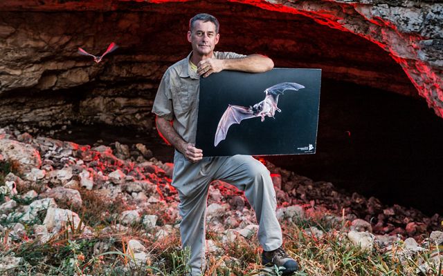 A man holds a photo of a bat outside the entrance of a cave