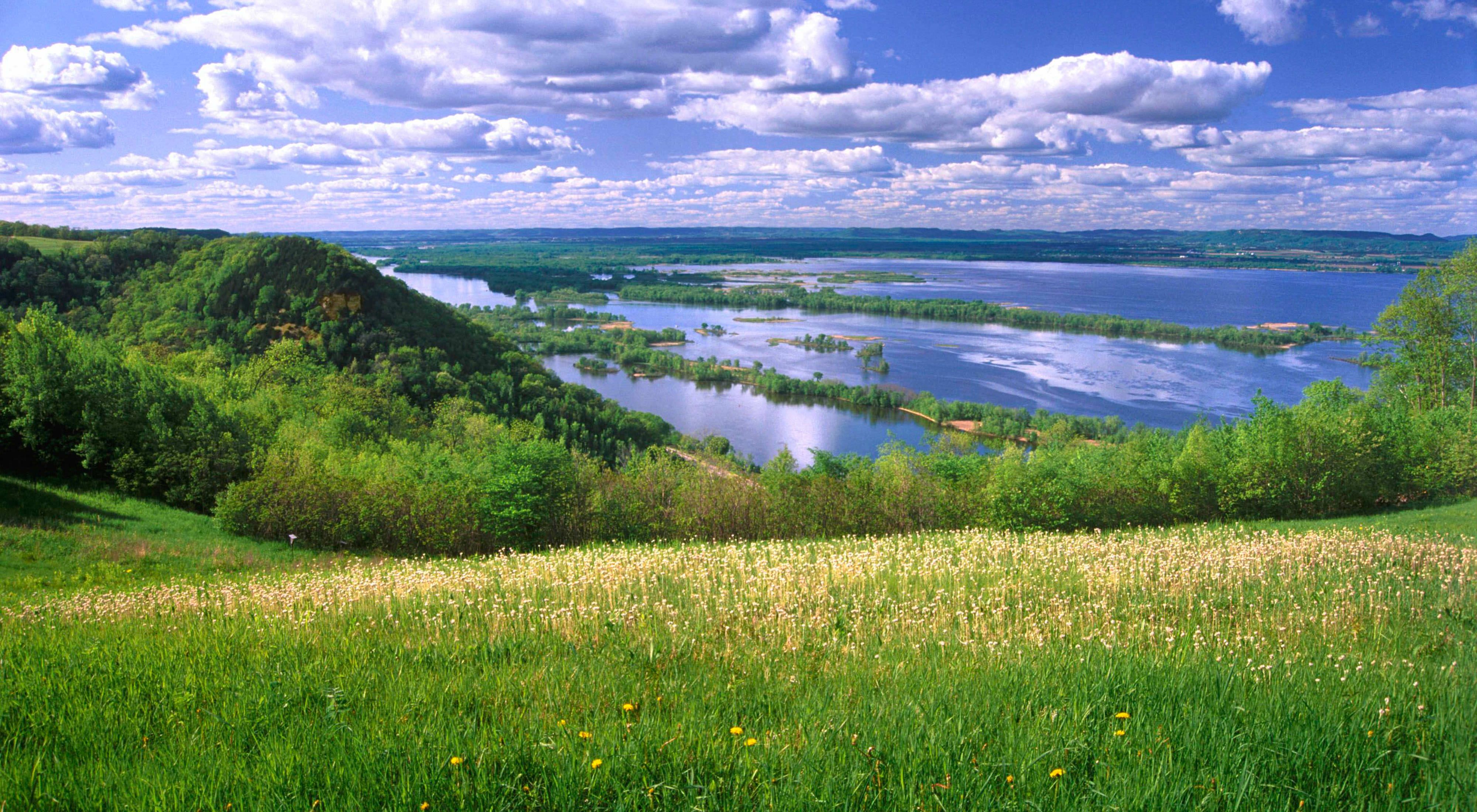 A green field overlooks a calm river and wetlands.