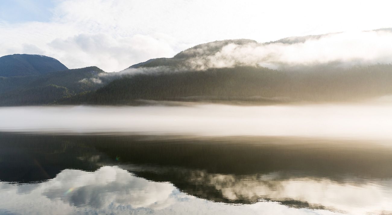 Hazy image of still water with forested mountain slopes in the background, partially shrouded by clouds.
