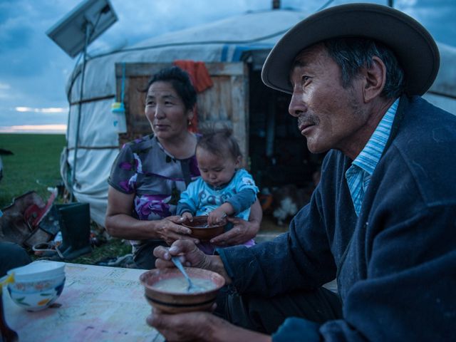 A man wearing a hat and a woman holding a child look into the distance.