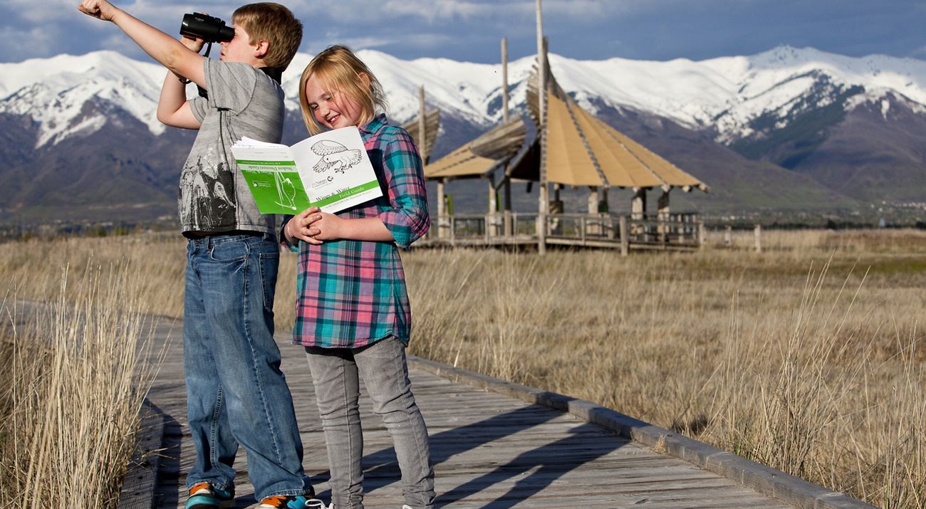 Two children smiling on a boardwalk with mountains and a building in the background.