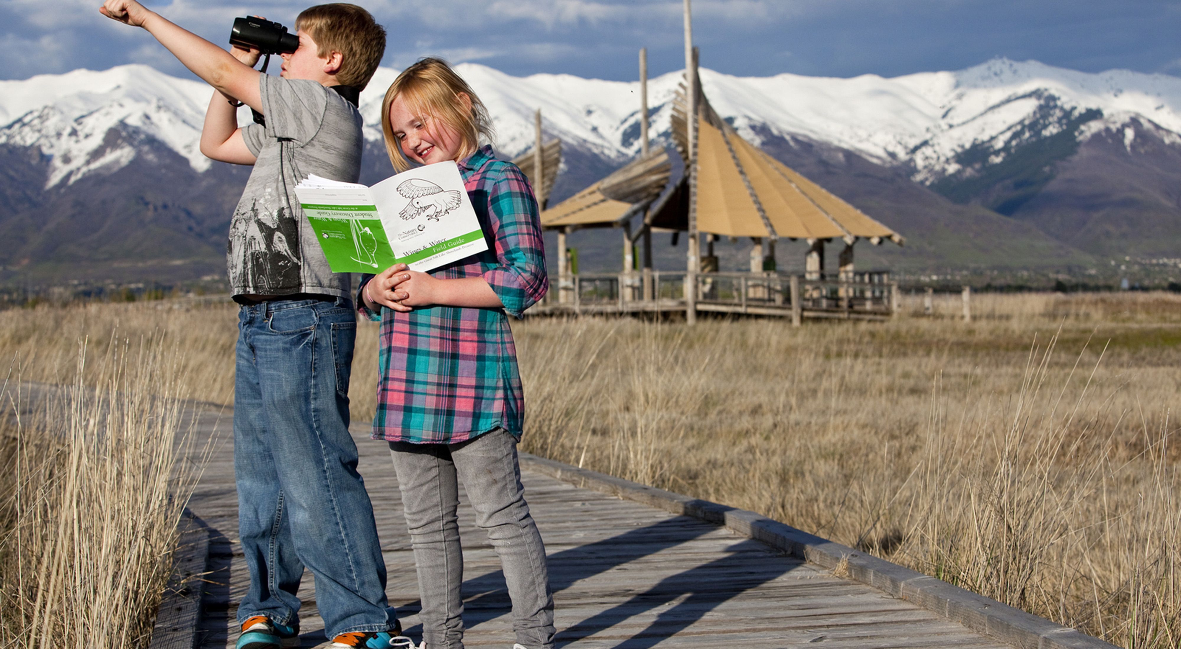 Two children smiling on a boardwalk with mountains and a building in the background. 